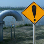 Tubería metálica cerca de un río con señal amarilla de advertencia, simbolizando riesgo de sanciones o incumplimiento normativo en sistemas de agua.